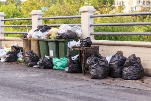 A collection of black and green rubbish bags, some partially open revealing tinted waste inside, stacked along a sidewalk next to a metal guardrail. The bags are placed directly on the pavement, with additional bags lined up along the base of the railing, indicating a waste collection or clearance activity. In the background, there is greenery with trees and a few buildings visible, suggesting an urban area near a park or residential zone in Lee. The scene is lit by natural daylight, providing clear visibility of the different textures and colors of the bags, from glossy black to matte green, and the rough surface of the pavement. The arrangement appears somewhat disorderly, typical of rubbish awaiting removal or disposal, and reflects typical rubbish accumulation seen during garden or waste clearance services in the Lee area. This scene is indicative of rubbish removal work carried out by Garden Clearance Lee, focusing on general waste collection from outdoor public or private spaces within the local vicinity.