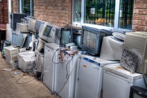 A row of discarded old computers and electronic devices arranged outdoors along a brick building wall, including CRT monitors, desktop towers, and fans in various sizes and colors, predominantly grey and black. Some cables and wires are visible, tangled and hanging from the devices. The equipment appears weathered with signs of dirt and dust accumulation, positioned on a paved surface adjacent to a window reflecting greenery. The scene depicts electronic waste that could be targeted for professional rubbish removal services, such as those offered by Garden Clearance Lee, to responsibly dispose of or recycle outdated technology in the Lee area, close to London. The background suggests an industrial or commercial setting, with the devices stacked minimally but in a somewhat cluttered manner. The overall atmosphere highlights proper management of electronic waste as part of garden clearance and rubbish removal tasks within the local community.