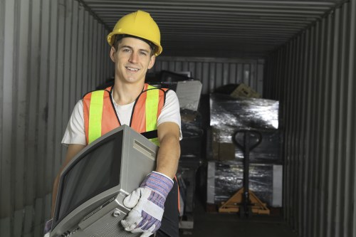 A young male worker wearing a yellow safety helmet, high-visibility safety vest with orange and yellow reflective stripes, and white work gloves is inside a metal storage container or shipping skip. He is holding a small, vintage-style television set with a curved screen and a plastic casing, which appears to be part of domestic rubbish. The container's interior features corrugated metal walls and is cluttered with wrapped or boxed items, including a pallet jack visible in the background. The environment suggests a waste disposal or clearance scenario, possibly linked to garden or household waste removal. The setting appears to be an outdoor or semi-covered area, with natural lighting illuminating the scene. This image is representative of rubbish removal services such as those provided by Garden Clearance Lee, focusing on the disposal of various household items and waste materials in and around Lee, South East London postcodes.
