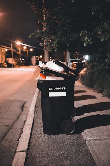 A black wheeled rubbish bin with the label 'ST. JOHN'S' is positioned on the edge of a paved sidewalk. The bin’s lid is partially open, revealing a mixture of paper, cardboard, and plastic waste insid