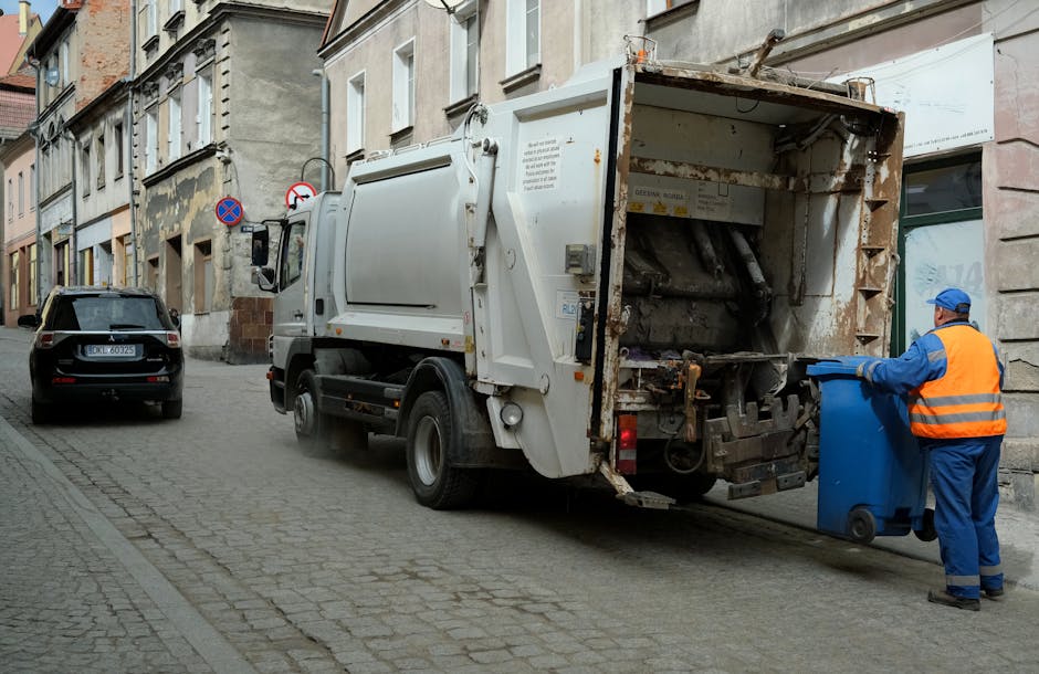 Two large black plastic garbage bags, filled and tightly tied at the top, are placed on the pavement next to a black metal fence. One bag appears slightly larger and is leaning against the other, with