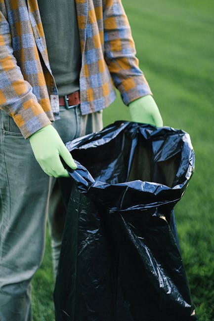 A person wearing a plaid shirt in shades of orange, gray, and blue, along with light green gloves, is holding open the top of a large black rubbish bag made of durable plastic, ready for waste disposa