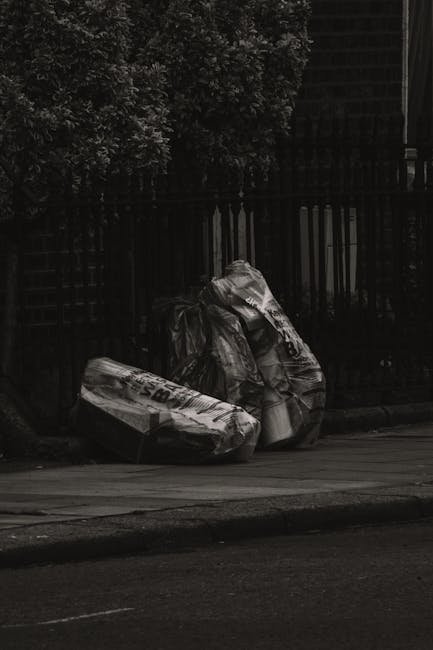 A person wearing a plaid shirt in shades of orange, gray, and blue, along with light green gloves, is holding open the top of a large black rubbish bag made of durable plastic, ready for waste disposa